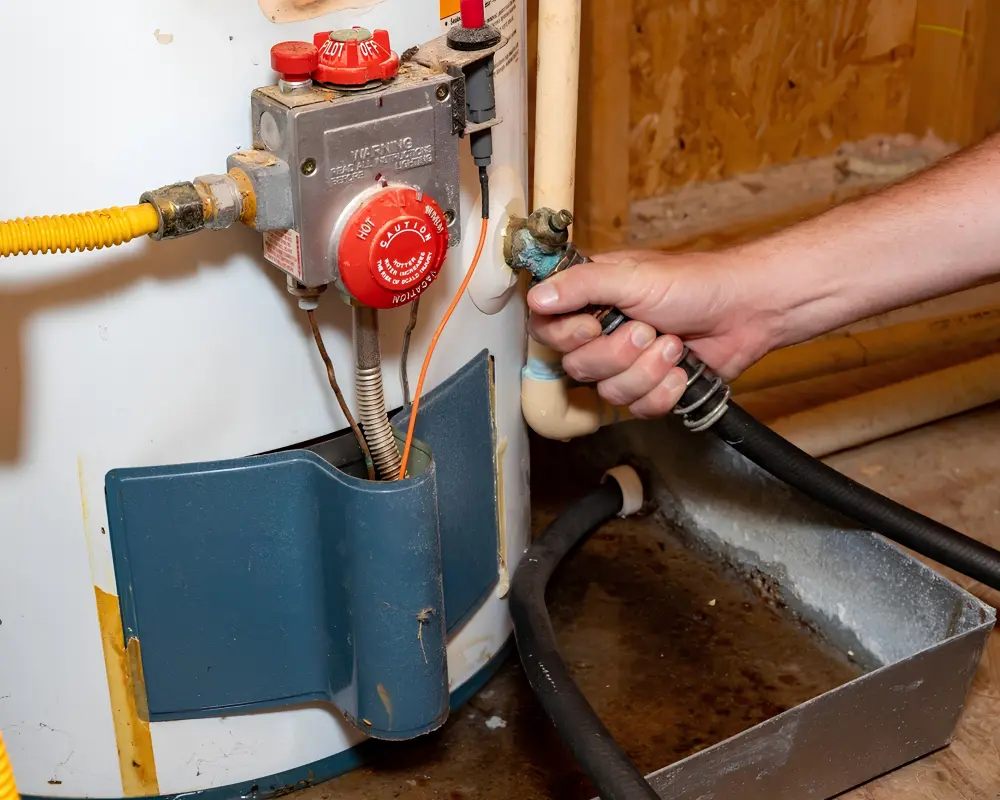 A close up view of a technician flushing a home hot water heater unit, connecting a black hose to the drain valve next to the gas control thermostat assembly during a water heater repair and maintenance service.